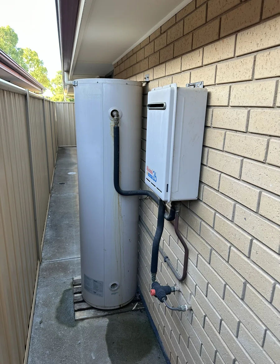 Old electric storage hot water tank and Rinnai gas unit mounted beside a brick wall before replacement